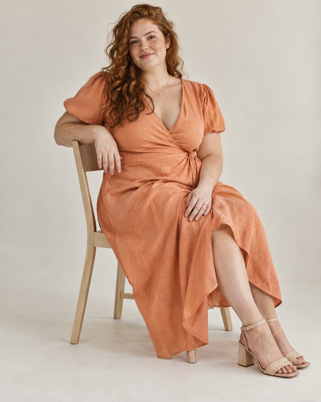 Adult plus-size woman with freckled fair skin and wavy auburn hair wearing an apricot cotton wrap midi dress, seated diagonally on a simple pale wooden chair in a cream photography studio