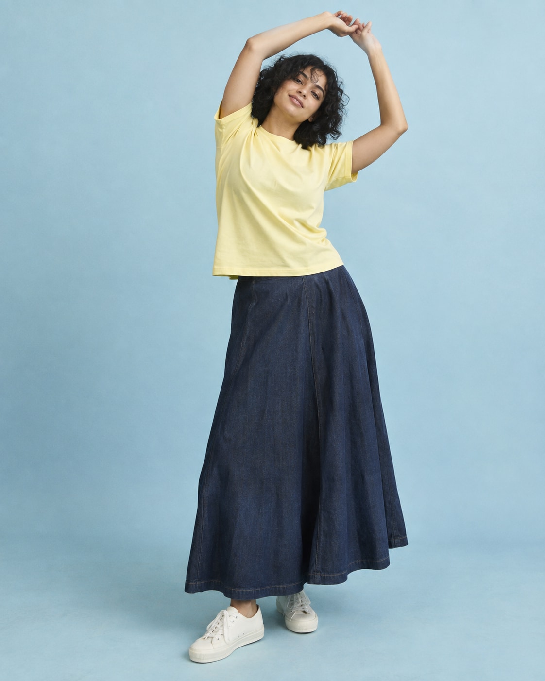 South Asian woman with curly bob in butter yellow cotton tee and long dark indigo skirt, arms in loose offset arc overhead and slight sideways lean, powder blue studio.