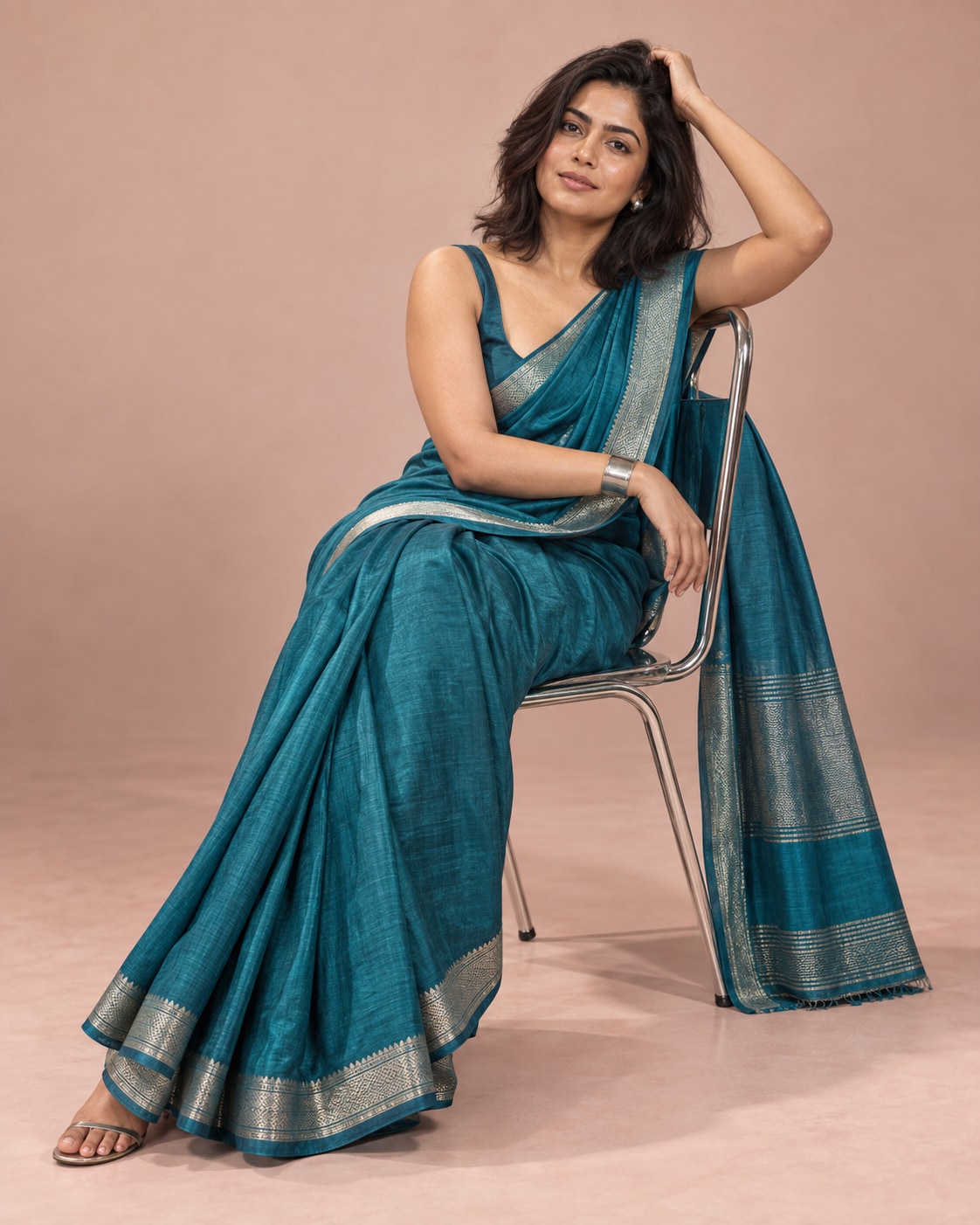 Indian woman in a turquoise silk saree seated sideways on a chrome chair, one arm curved above her head against a warm blush studio backdrop.