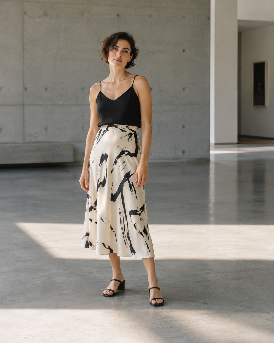 An adult short-haired woman wearing black camisole and cream abstract-print midi skirt, full length standing in an airy concrete gallery, generous architectural negative space and a rectangle of light on the floor, soft luminous face.