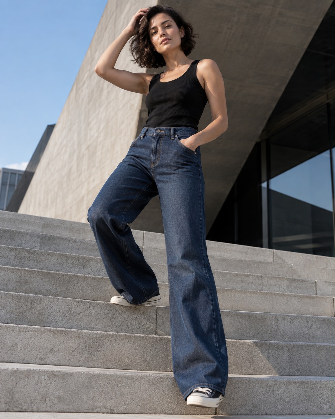 Adult woman with short dark hair wearing black ribbed tank and high-waisted dark blue wide jeans, standing on broad modern concrete steps with one foot one step higher