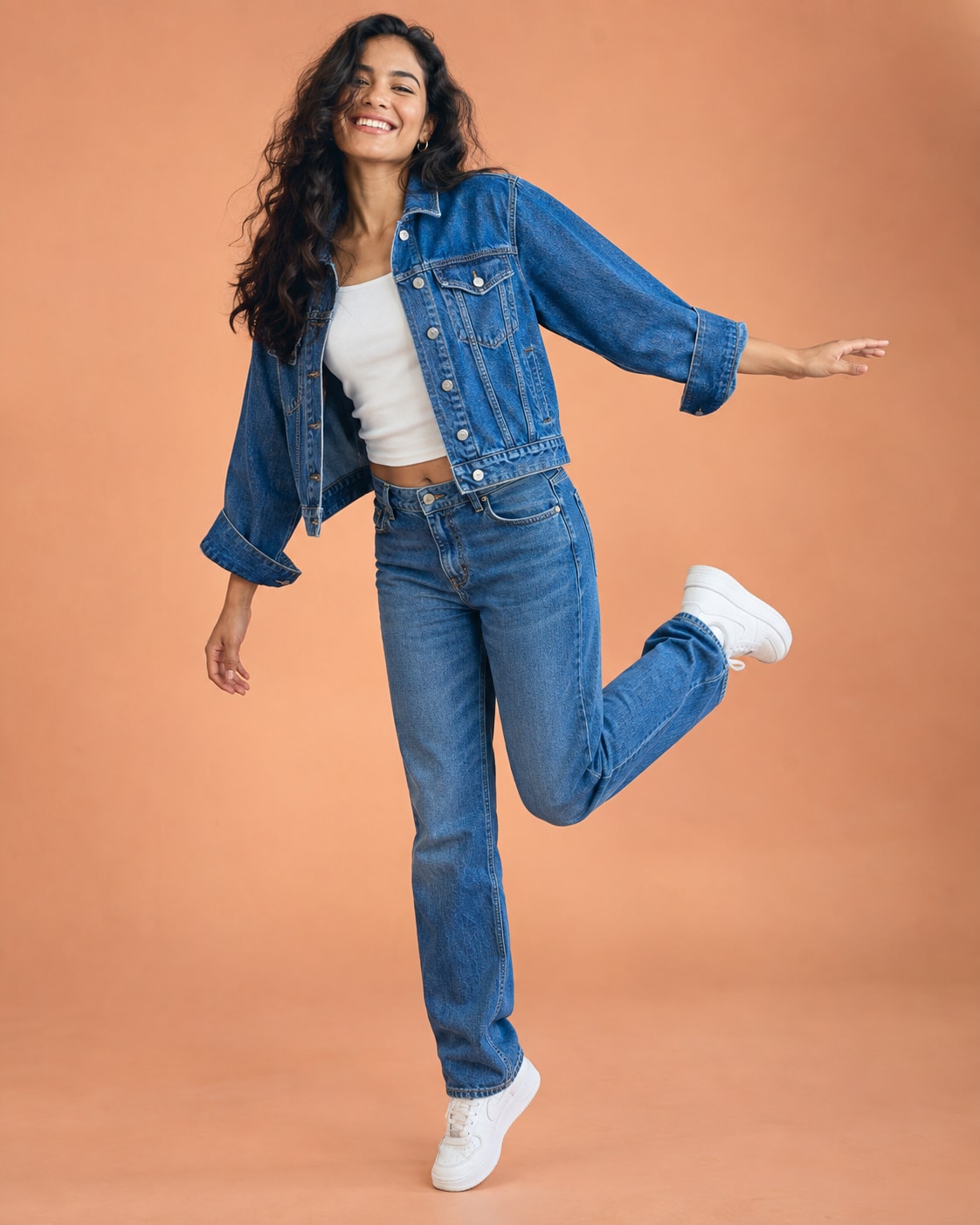 Latina woman in cobalt denim jacket and straight jeans, white sneakers, balancing on one leg with other bent back and one arm extended, full body on peach studio sweep.