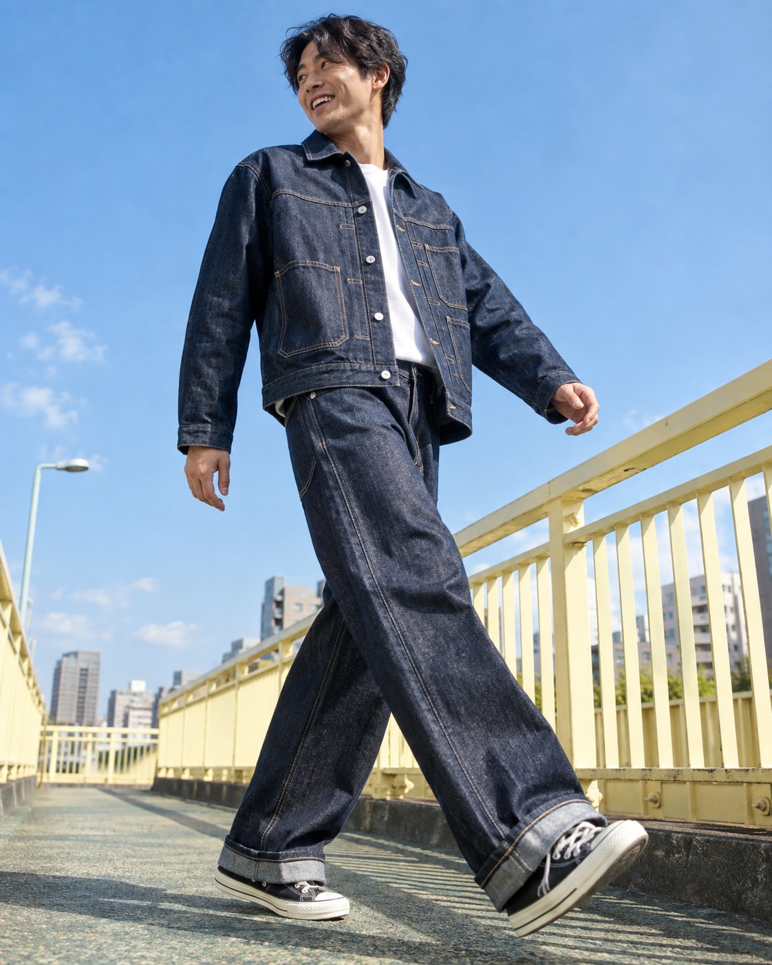 East Asian man in indigo denim co-ord taking exaggerated natural long stride on pedestrian overpass with pale yellow railings, arms swinging, laughing sideways glance, low angle and bright blue sky.