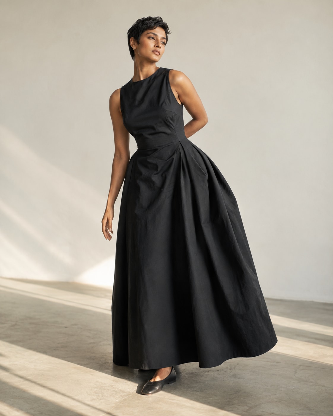 Brown-skinned woman with pixie hair in sculptural black cotton maxi dress, twisting gently at waist with one arm behind and one relaxed, broad natural skirt curve, cream studio and diagonal floor shadow.