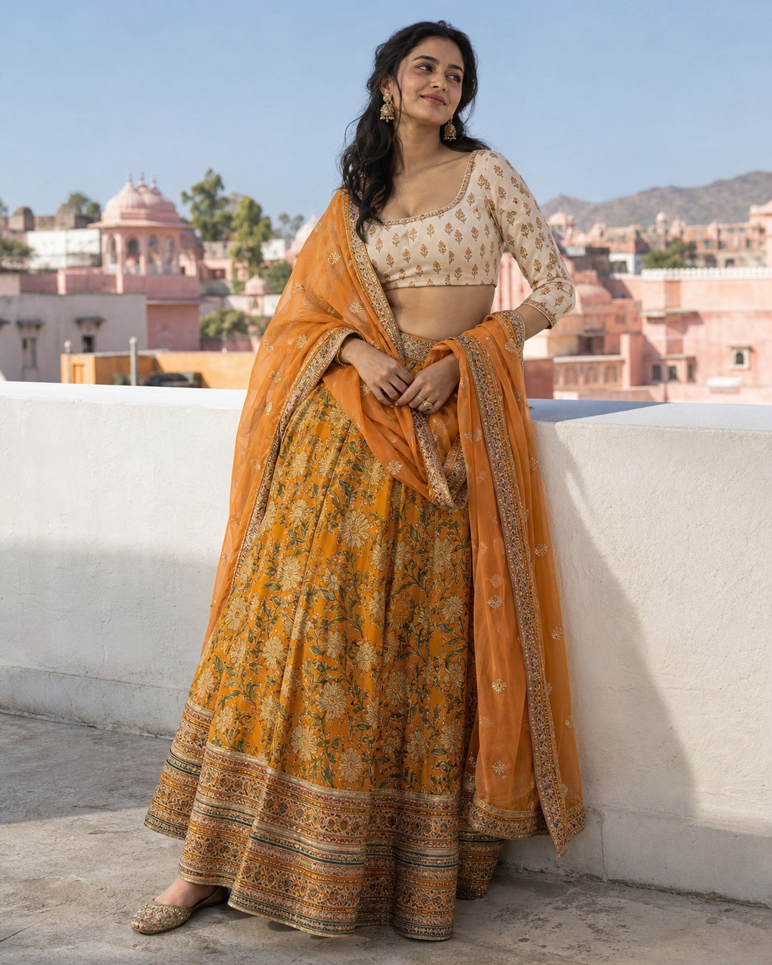 South Asian woman in marigold block-print lehenga, cream blouse and sheer orange dupatta leaning hip lightly at broad white rooftop parapet, hands low holding dupatta edges, pink city roofs and clear sky.