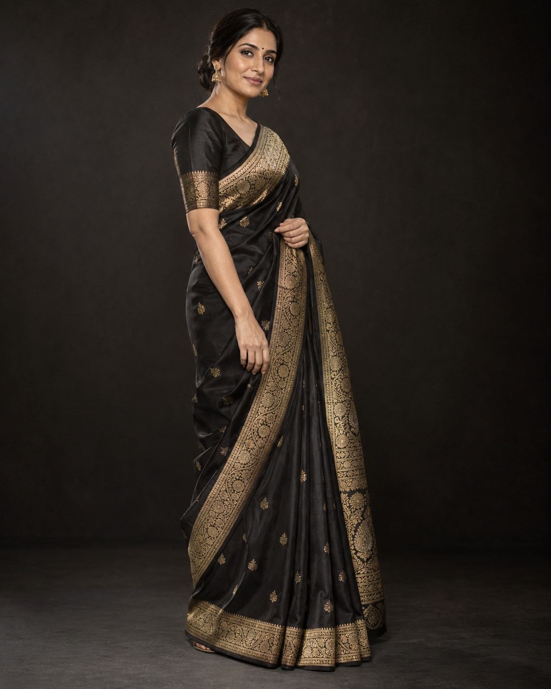 Indian woman in a black silk saree with antique-gold zari standing against a deep charcoal backdrop, her face and woven border softly illuminated.