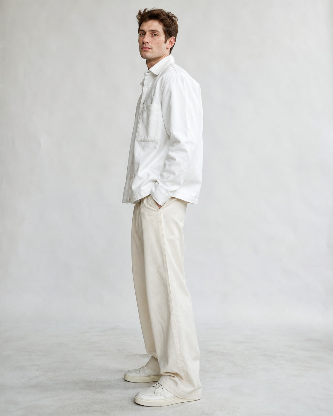 Freckled white man in crisp white boxy overshirt and ecru trousers, standing side profile with face turned gently to camera, one hand in pocket, raw white paper backdrop.