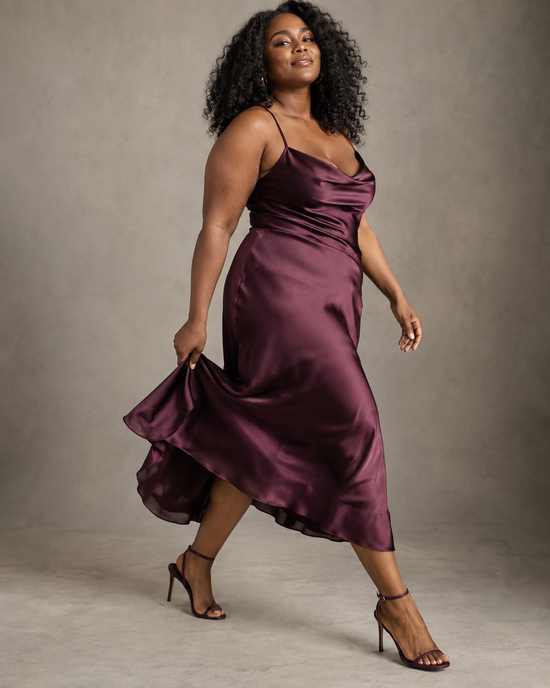 Curvy Black woman in deep plum satin midi dress taking broad sideways step, holding a little edge of skirt so it sweeps naturally, warm grey studio and confident expression.