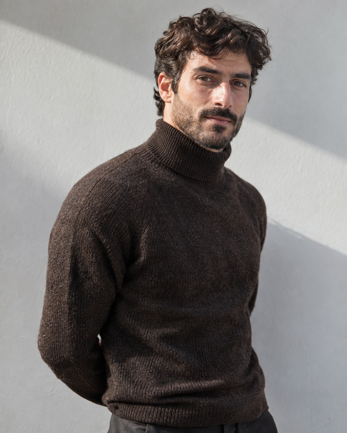 Model in a dark knitted turtleneck, turning one shoulder towards the camera against a grey studio wall crossed by diagonal light.
