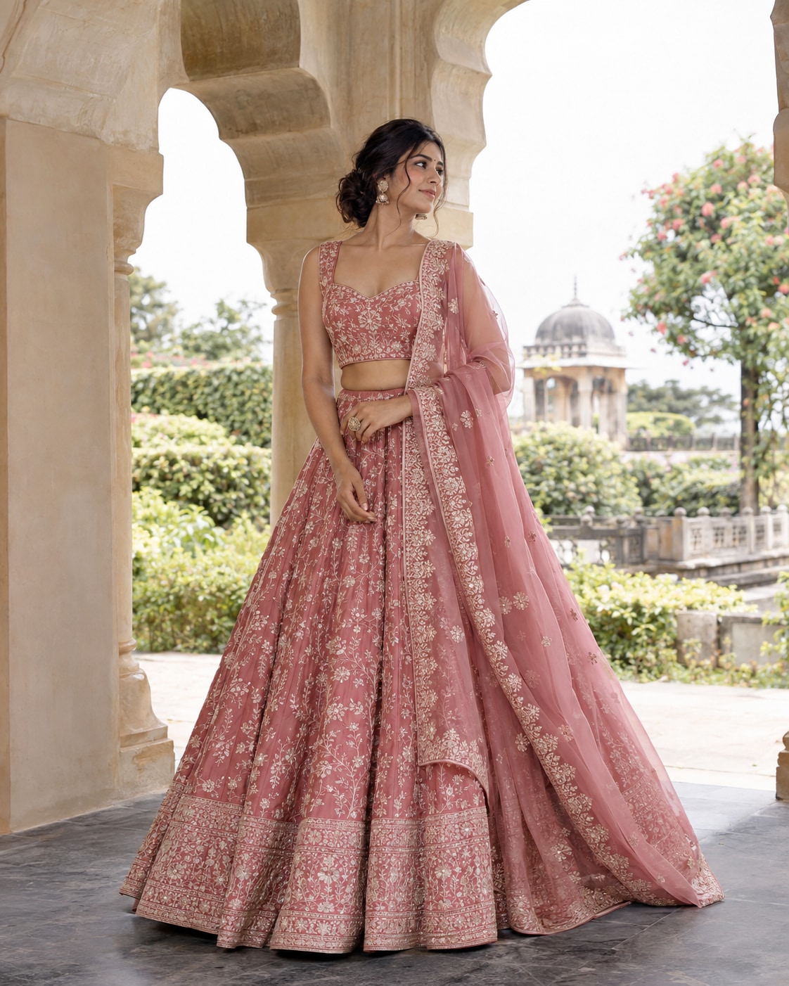 Adult Indian woman wearing a rose-pink embroidered lehenga with fine ivory floral threadwork and matching sheer dupatta, standing in a bright historic garden beneath a pale stone arch