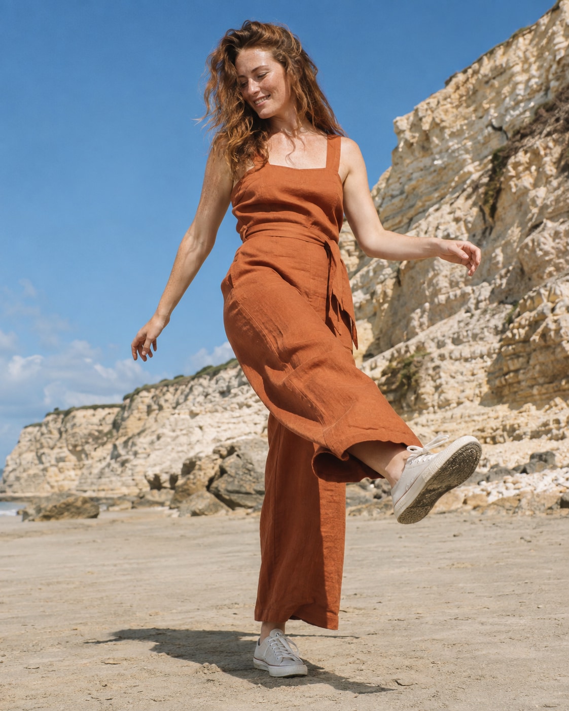 Freckled white woman in burnt orange linen jumpsuit doing small playful forward kick with arms loosely out, level sand by pale sandstone cliffs, full body and bright blue sky.
