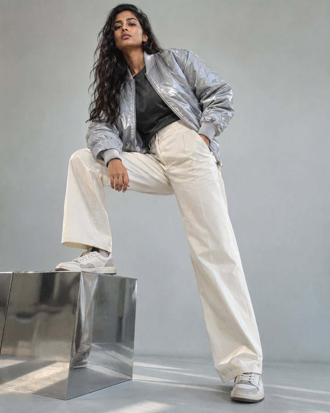 South Asian woman in silver lightweight bomber, charcoal tee and cream trousers, stepping onto low chrome platform with bent knee, pale grey studio, low camera angle, realistic reflective nylon.