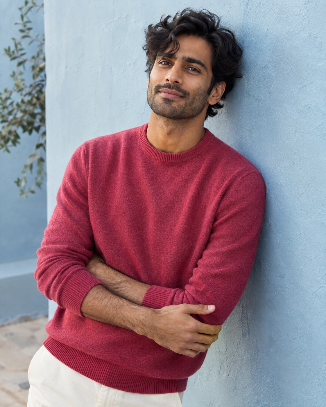 South Asian man with wavy hair in raspberry fine knit crewneck leaning one shoulder against pale blue plaster wall, arms folded loosely low, waist-up intimate relaxed portrait.