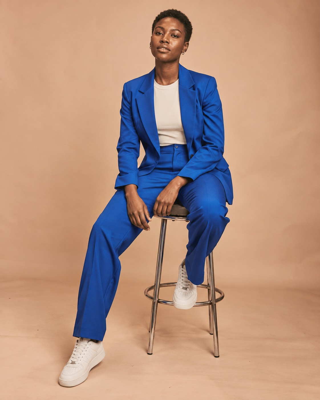 Adult Black woman with close-cropped natural hair in vivid cobalt blue tailored suit and cream tee, sitting sideways on simple chrome stool in a warm pale peach photography studio