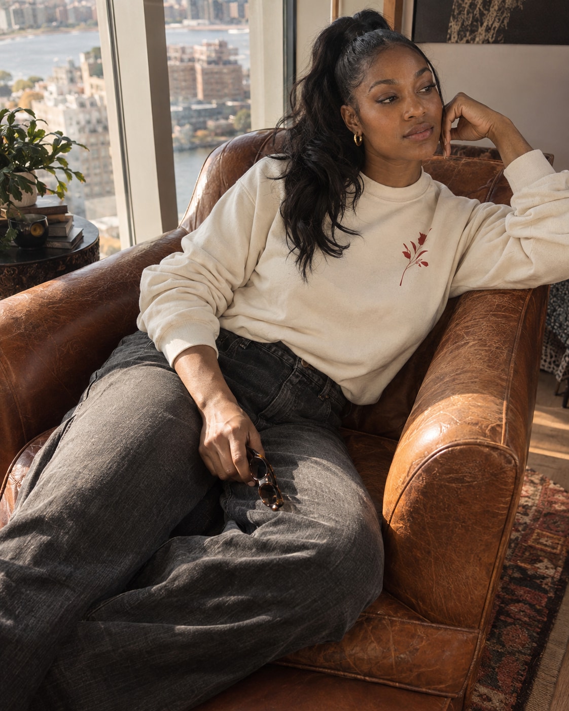 An adult Black woman with long wavy ponytail wearing a cream sweatshirt with a small original abstract red botanical graphic and charcoal jeans lounges diagonally in a cognac leather armchair by a tall apartment window